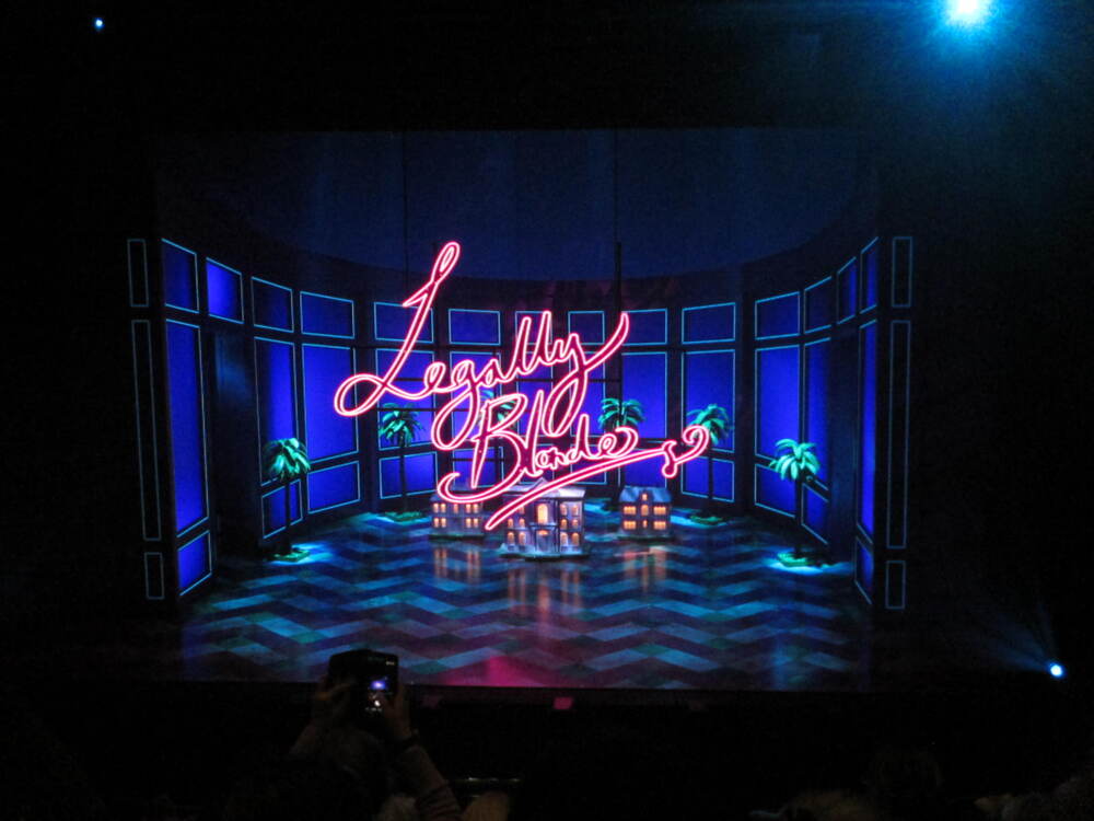 A blue-lit modern stage with a large pink neon sign in front that says "Legally Blonde" in cursive