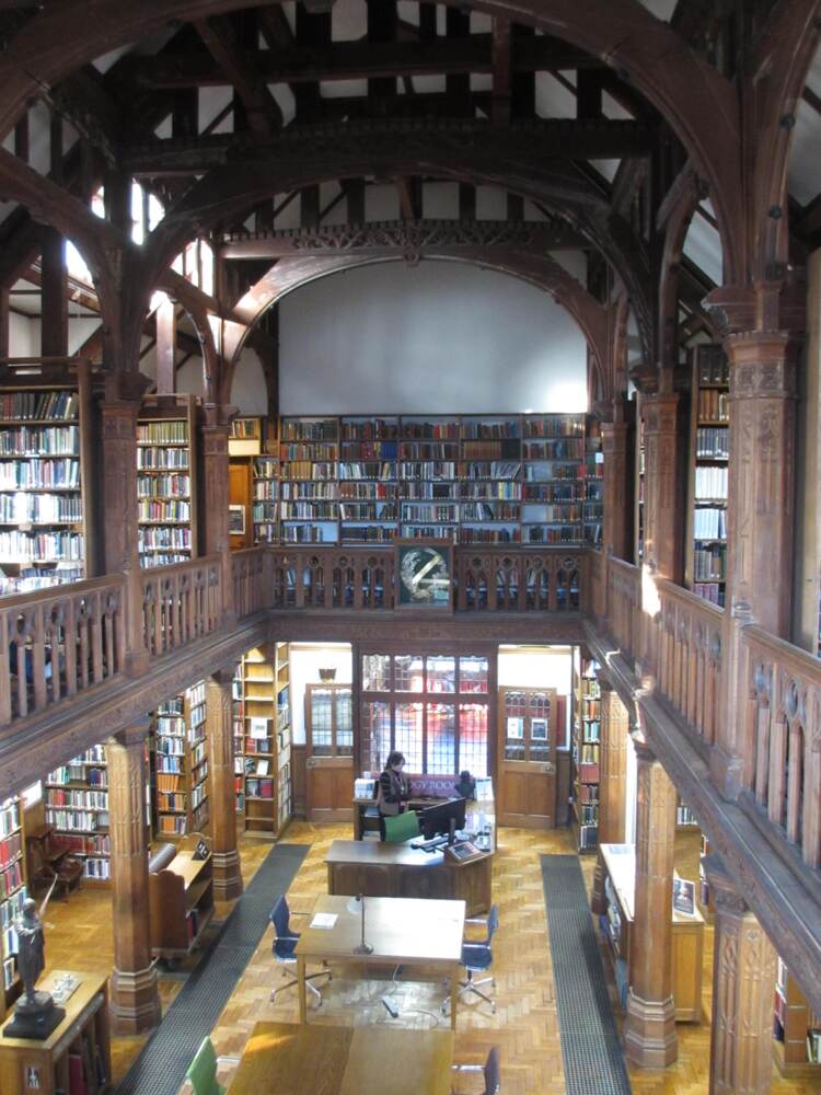 a beuatiful reading room with white walls, brown wood bookshelves full of old books, brown arches in the ceiling and daylight streaming in from non-visible walls on the top of two floors. the bottom floor, visible through the middle over the balcony, is warmly lit with armchairs.